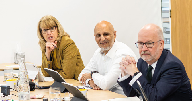 Board members in a meeting — three directors seated at a table with laptops and water bottles, engaged in discussion